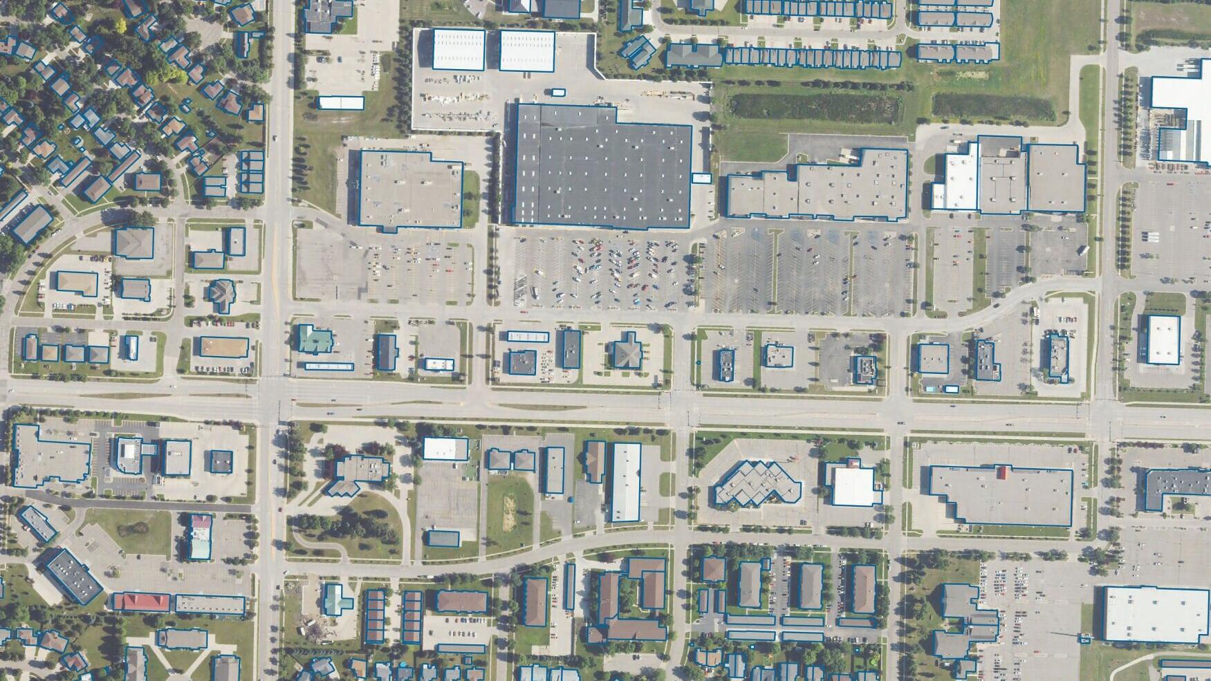 This image shows building footprints detected by EarthDefine's Buildings Footprints 3D API, outlined in blue over aerial imagery of a mixed-use district. The API identifies individual structures across residential, commercial, and industrial areas natio...