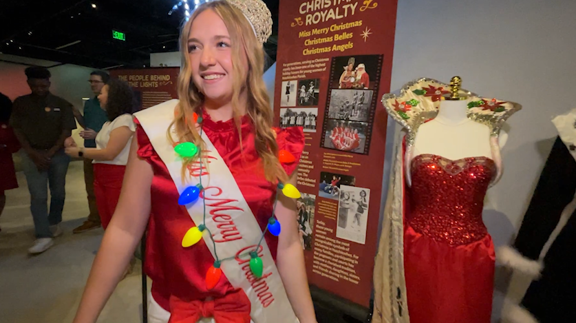 Reigning Miss Merry Christmas in front of display honoring 7 decades of pageantry