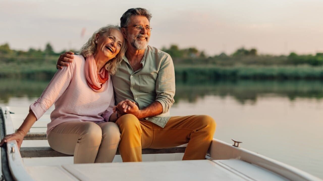 Elderly couple holding hands and laughing, enjoying a peaceful moment together while boating on a calm lake at sunset, embracing retirement and sharing a romantic experience