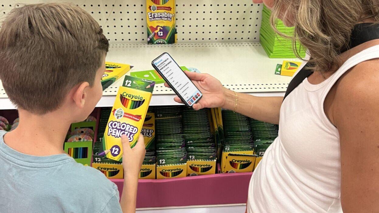 A mother uses TeacherLists on her phone while her son selects back-to-school supplies from his list.