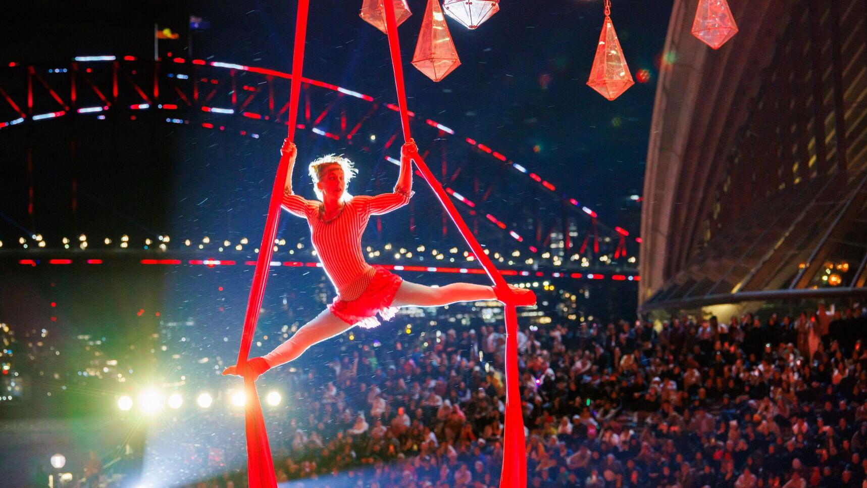 An aerial performer in a red costume executes a split on red silks during a Vivid Sydney 2026 event, with the Sydney Harbour Bridge and Opera House illuminated in the background and a large audience watching