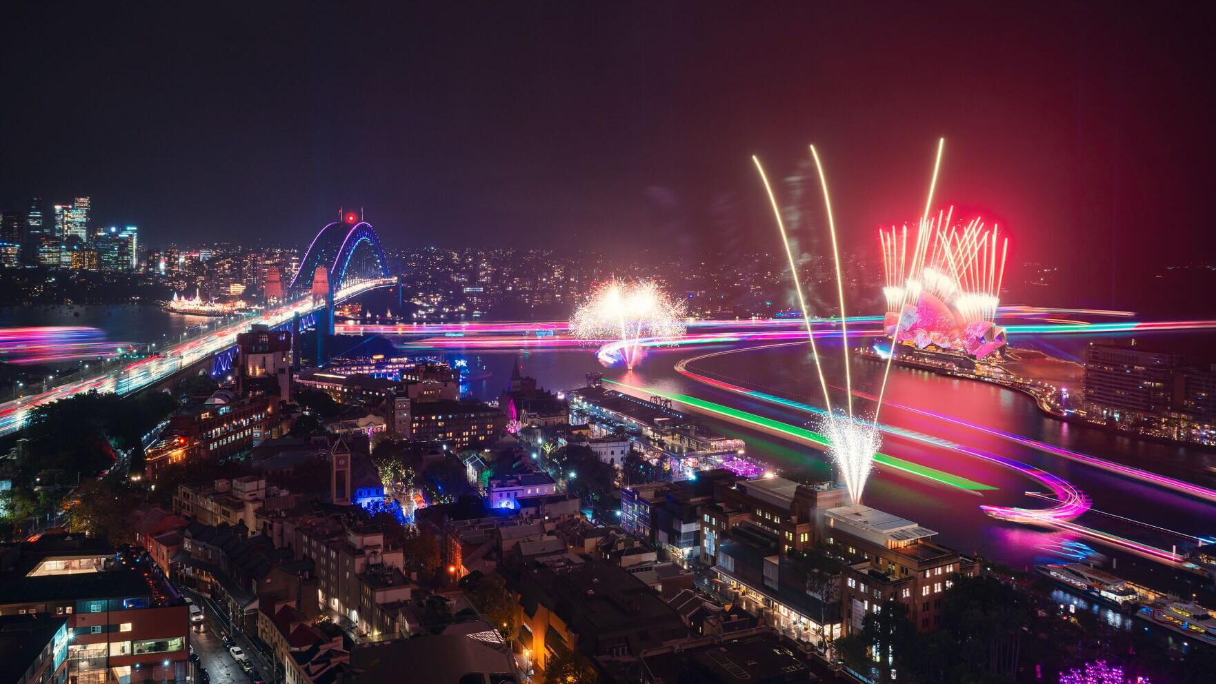 A panoramic night view of Sydney showcasing Vivid Sydney 2026, with fireworks illuminating the sky above the Sydney Opera House and Harbour Bridge